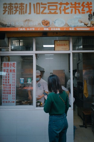 A woman is standing outside a small food stall, looking at her phone. Inside the stall, a person wearing a mask and a chef's hat is preparing food. There is a menu displayed on the window, and a large sign with Chinese characters is above the stall.