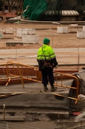A construction worker wearing a bright yellow-green safety jacket and a green hat stands on a construction site with orange barriers. Surrounding the worker are various construction materials and partially built structures. The background includes a large structure covered in green tarp.