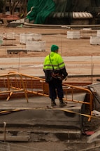 A construction worker wearing a bright yellow-green safety jacket and a green hat stands on a construction site with orange barriers. Surrounding the worker are various construction materials and partially built structures. The background includes a large structure covered in green tarp.