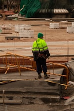 A construction worker wearing a bright yellow-green safety jacket and a green hat stands on a construction site with orange barriers. Surrounding the worker are various construction materials and partially built structures. The background includes a large structure covered in green tarp.