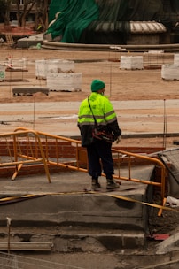 A construction worker wearing a bright yellow-green safety jacket and a green hat stands on a construction site with orange barriers. Surrounding the worker are various construction materials and partially built structures. The background includes a large structure covered in green tarp.