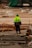 A construction worker wearing a bright yellow-green safety jacket and a green hat stands on a construction site with orange barriers. Surrounding the worker are various construction materials and partially built structures. The background includes a large structure covered in green tarp.