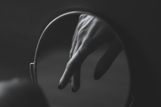 A close-up view of a hand with long fingers and painted nails, reflected in a circular mirror against a dark background.