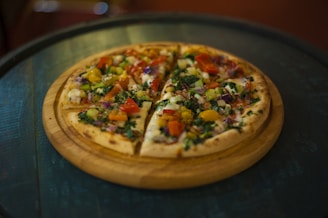 A freshly baked vegetable pizza is presented on a round wooden serving board. The pizza is generously topped with a colorful assortment of ingredients including tomatoes, bell peppers, onions, broccoli, and melted cheese. The crust appears crispy and golden brown, providing a contrasting texture to the vibrant toppings.