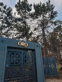 A blue metal gate with intricate geometric patterns stands in front of tall trees with dense foliage. The top of the gate features a sign with the word 'ORO' in gold lettering. The background is filled with the branches and leaves of the trees, creating a natural, outdoor setting.