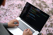 Close-up of hands typing code on a laptop with a backdrop of cornfields visible through a window.