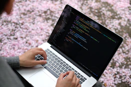 Close-up of hands typing code on a laptop with a backdrop of cornfields visible through a window.