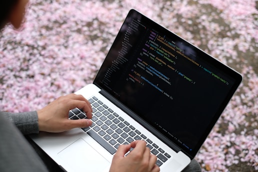 A person is typing on a laptop keyboard, which displays a code editor with various lines of colorful programming code against a black background. The hands are positioned over the keyboard, indicating active typing or coding. In the background, pink and white petals are scattered, suggesting a natural and calm setting.