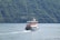A ferry glides through calm waters, creating waves behind it. The vessel has two decks with seating areas, and is adorned with red trim accents. Dense green hills surround the body of water, with scattered small houses along the hillside.