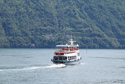 A ferry glides through calm waters, creating waves behind it. The vessel has two decks with seating areas, and is adorned with red trim accents. Dense green hills surround the body of water, with scattered small houses along the hillside.