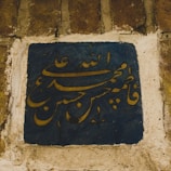 Arabic calligraphy with elegantly curved letters on a deep blue ceramic tile, set into a weathered stone wall.