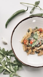 A plate of fusilli pasta garnished with fresh basil and topped with cherry tomatoes and grated cheese. Surrounding the plate are fresh green herbs, a garlic clove, and a green chili pepper, all laid out on a white surface.