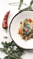 A plate of fusilli pasta garnished with cherry tomatoes, fresh basil leaves, and grated cheese is artfully arranged on a white surface. Next to the plate, there is a red chili pepper, a clove of garlic, and sprigs of green basil, creating a fresh and inviting presentation.
