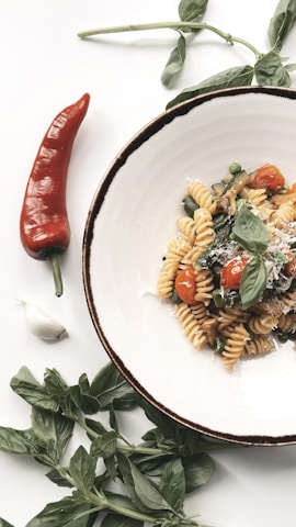 A plate of fusilli pasta garnished with cherry tomatoes, fresh basil leaves, and grated cheese is artfully arranged on a white surface. Next to the plate, there is a red chili pepper, a clove of garlic, and sprigs of green basil, creating a fresh and inviting presentation.