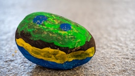 A small rock painted with vibrant colors, featuring green on the top, a black band with yellow in the middle, and blue on the bottom. Two blue circular patterns are visible on the top green section. The rock rests on a textured, speckled gray surface.