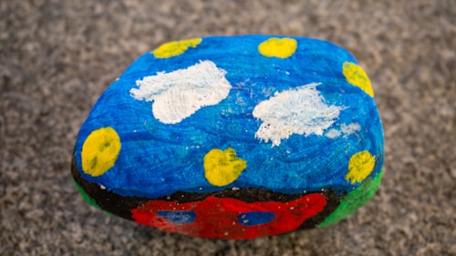 A rock painted with a bright blue background featuring white cloud-like shapes and scattered yellow circles. The lower part of the rock displays contrasting colors, including red, black, and green. The surface underneath the rock appears to be a rough gray texture.