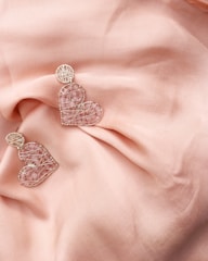 Two heart-shaped earrings made of intricate wire and adorned with small pink stones rest on a soft, peach-colored fabric. The earrings have a delicate and artistic design, with a circular element attached to each heart. The fabric is gently draped, adding a touch of elegance and softness to the overall composition.