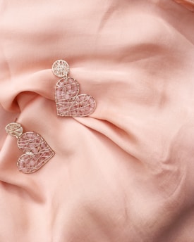 Two heart-shaped earrings made of intricate wire and adorned with small pink stones rest on a soft, peach-colored fabric. The earrings have a delicate and artistic design, with a circular element attached to each heart. The fabric is gently draped, adding a touch of elegance and softness to the overall composition.