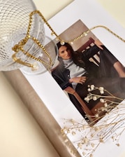 A stylish magazine layout featuring a woman's portrait is adorned with a delicate gold chain and glass cup. The scene is complemented by small white flowers lying on the open page, creating an artistic and elegant arrangement.