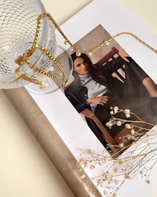 A stylish magazine layout featuring a woman's portrait is adorned with a delicate gold chain and glass cup. The scene is complemented by small white flowers lying on the open page, creating an artistic and elegant arrangement.