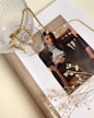 A stylish magazine layout featuring a woman's portrait is adorned with a delicate gold chain and glass cup. The scene is complemented by small white flowers lying on the open page, creating an artistic and elegant arrangement.