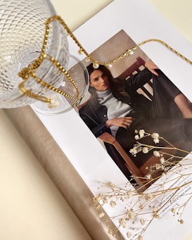 A stylish magazine layout featuring a woman's portrait is adorned with a delicate gold chain and glass cup. The scene is complemented by small white flowers lying on the open page, creating an artistic and elegant arrangement.