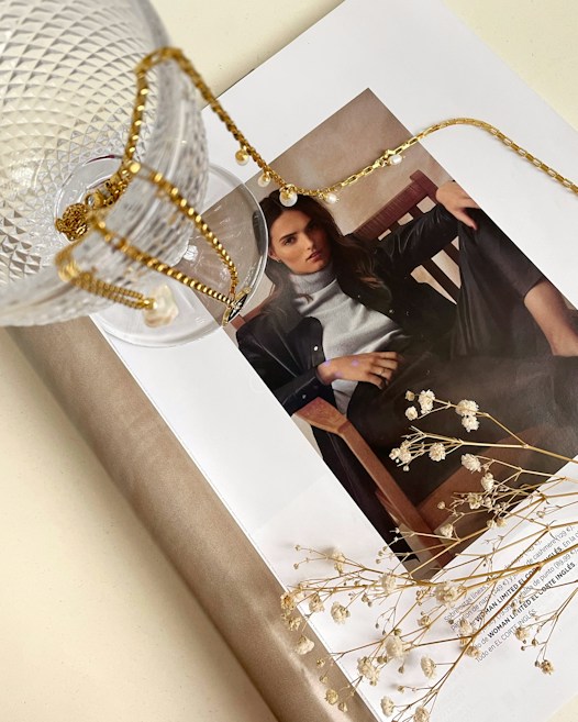 A stylish magazine layout featuring a woman's portrait is adorned with a delicate gold chain and glass cup. The scene is complemented by small white flowers lying on the open page, creating an artistic and elegant arrangement.