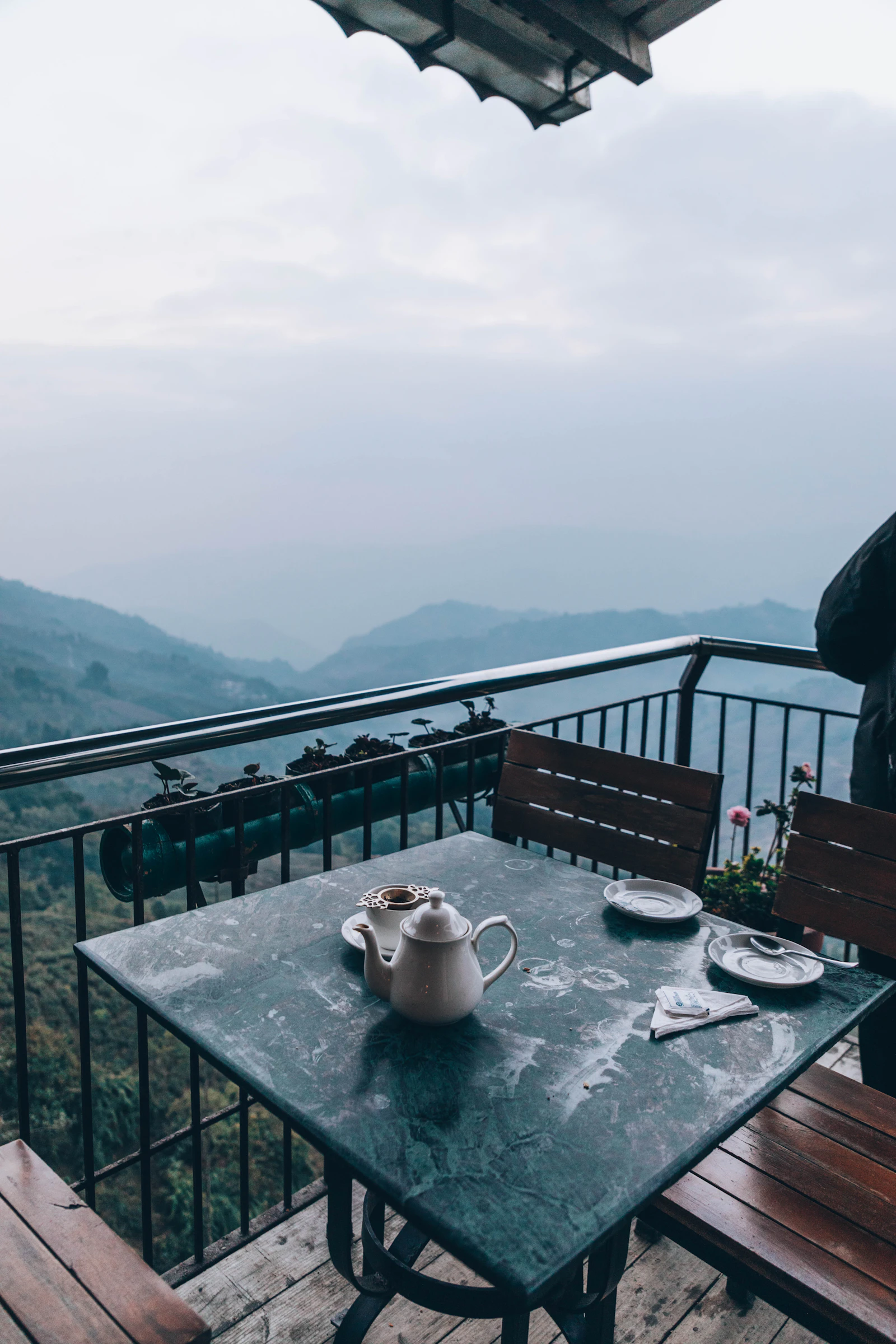 A teapot and two cups on a balcony table in Darjeeling, green hills fading into haze beyond the railing