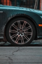 A close-up view of a car's wheel with a shiny alloy rim, partially covered with road dirt. The car body is dark with some visible grime and the background shows a cracked pavement. There is a warm orange reflection in the background, suggesting urban surroundings.