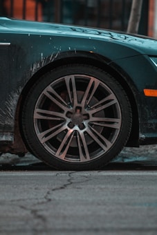 A close-up view of a car's wheel with a shiny alloy rim, partially covered with road dirt. The car body is dark with some visible grime and the background shows a cracked pavement. There is a warm orange reflection in the background, suggesting urban surroundings.