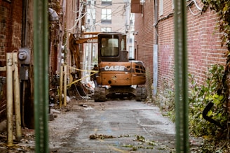 A narrow alleyway lined with red brick walls features a small excavator with the brand name 'CASE' visible. The alley is cluttered with debris, caution tape, and overgrown weeds, giving a sense of ongoing construction. Utility lines and pipes run along the walls, and the ground shows signs of recent work.