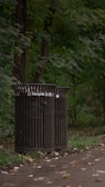 A recycling bin is positioned on a forested pathway, surrounded by lush green trees and scattered fallen leaves. The ground is a paved path lined with grass on the edges.
