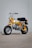 A small, yellow and white mini bike is parked on a concrete surface with a plain gray wall in the background. It features a retro design with a single headlight, black seat, and chunky tires.