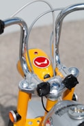 Close-up view of the handlebars and headlight area of a bright yellow bicycle. The metal handlebars are shiny and chrome-plated, while the casing around the headlight features a red logo. The structure is accentuated by bolts and cables, showcasing a mixture of modern and retro design elements.
