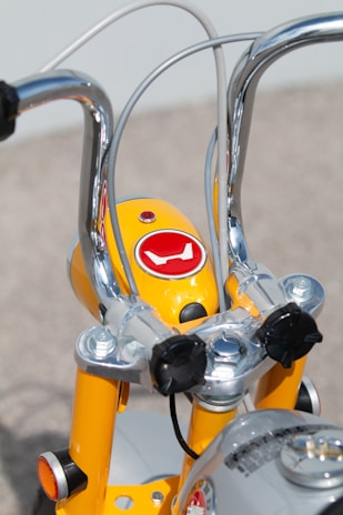 Close-up view of the handlebars and headlight area of a bright yellow bicycle. The metal handlebars are shiny and chrome-plated, while the casing around the headlight features a red logo. The structure is accentuated by bolts and cables, showcasing a mixture of modern and retro design elements.