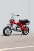 A red Honda minibike is parked on a concrete surface against a plain light grey wall. The bike features a prominent black seat, chrome handlebars, and large knobby tires. The overall style is vintage, with shiny chrome accents and a compact design.