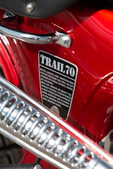 Close-up view of a red, polished metallic surface of a vehicle with a label reading 'TRAIL 70'. The label contains safety and operation instructions. There is a shiny metal handle and a part of the exhaust system visible in the foreground.