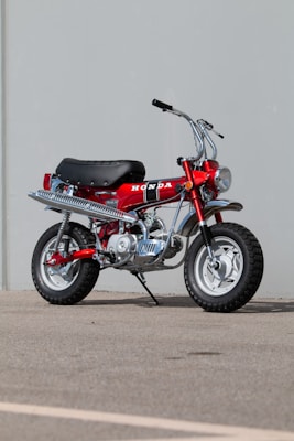 A classic Honda mini bike with a bright red frame, black seat, and large knobby tires. The motorcycle features silver exhaust and chrome details, parked on a paved surface against a plain, light grey background.