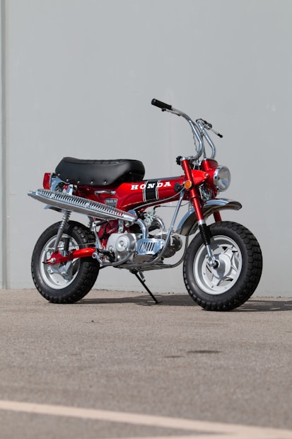 A classic Honda mini bike with a bright red frame, black seat, and large knobby tires. The motorcycle features silver exhaust and chrome details, parked on a paved surface against a plain, light grey background.