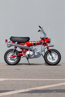 A vintage red motorcycle with a black seat and chrome accents is parked on a concrete surface against a plain gray backdrop. The motorcycle features a prominent Honda logo on the side, along with knobby tires designed for off-road use.