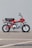 A vintage red motorcycle with a black seat and chrome accents is parked on a concrete surface against a plain gray backdrop. The motorcycle features a prominent Honda logo on the side, along with knobby tires designed for off-road use.