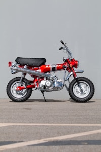A vintage red motorcycle with a black seat and chrome accents is parked on a concrete surface against a plain gray backdrop. The motorcycle features a prominent Honda logo on the side, along with knobby tires designed for off-road use.