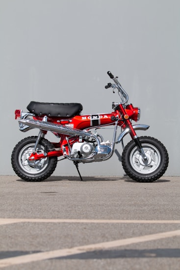 A vintage red motorcycle with a black seat and chrome accents is parked on a concrete surface against a plain gray backdrop. The motorcycle features a prominent Honda logo on the side, along with knobby tires designed for off-road use.