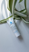 A small white tube of skincare product lies on a light background, with large green and white striped leaves partially draped over it. The tube has blue and black text printed on it.