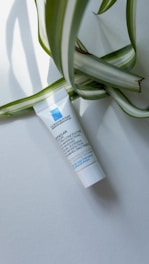 A small white tube of skincare product lies on a light background, with large green and white striped leaves partially draped over it. The tube has blue and black text printed on it.