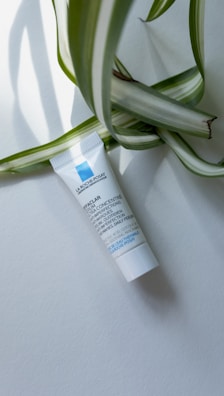 A small white tube of skincare product lies on a light background, with large green and white striped leaves partially draped over it. The tube has blue and black text printed on it.