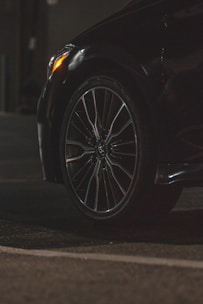 A close-up view of a car tire and wheel, featuring a sleek alloy rim. The surroundings are dimly lit, highlighting the reflective surface of the tire and part of the car body. The foreground is asphalt, suggesting the car is parked or in a driveway.