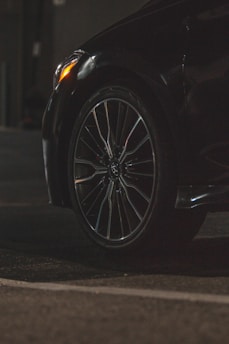 A close-up view of a car tire and wheel, featuring a sleek alloy rim. The surroundings are dimly lit, highlighting the reflective surface of the tire and part of the car body. The foreground is asphalt, suggesting the car is parked or in a driveway.