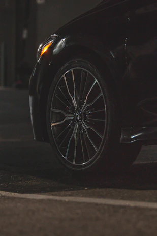 A close-up view of a car tire and wheel, featuring a sleek alloy rim. The surroundings are dimly lit, highlighting the reflective surface of the tire and part of the car body. The foreground is asphalt, suggesting the car is parked or in a driveway.