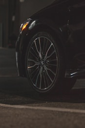 A close-up view of a car tire and wheel, featuring a sleek alloy rim. The surroundings are dimly lit, highlighting the reflective surface of the tire and part of the car body. The foreground is asphalt, suggesting the car is parked or in a driveway.